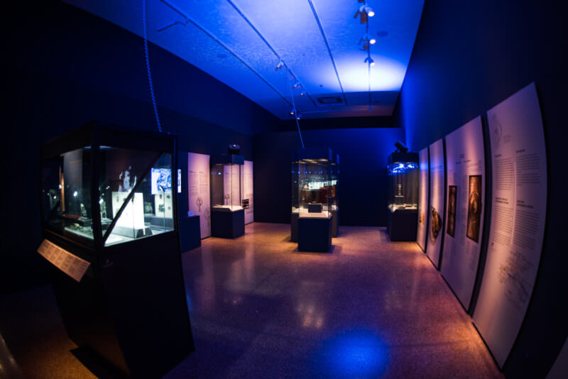 Antikythera Shipwreck Exhibition gallery with blue ceiling lighting, glass display cases, and illuminated text panels along the walls