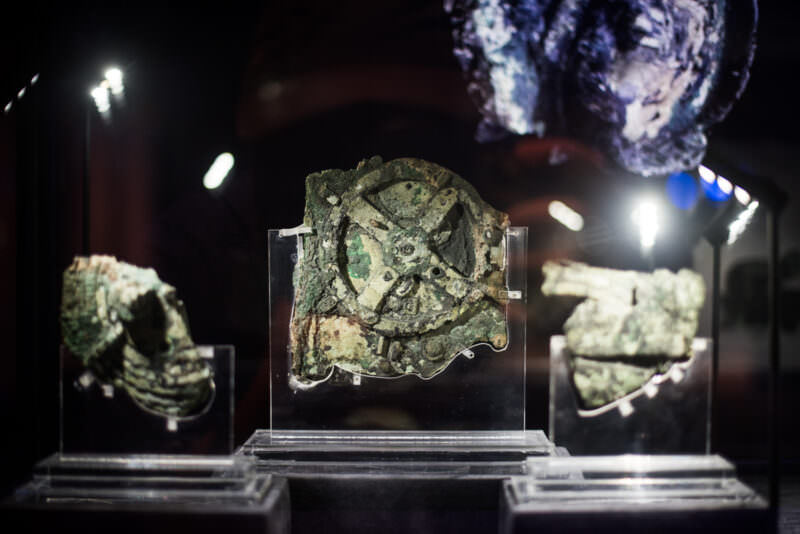 Antikythera Mechanism Fragment A in clear case, four-spoked main gear prominently visible in corroded bronze with green and brown patina