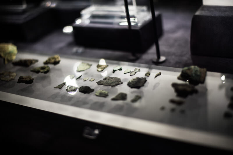 Small corroded bronze fragments of the Antikythera Mechanism scattered on a white display surface, shallow depth of field