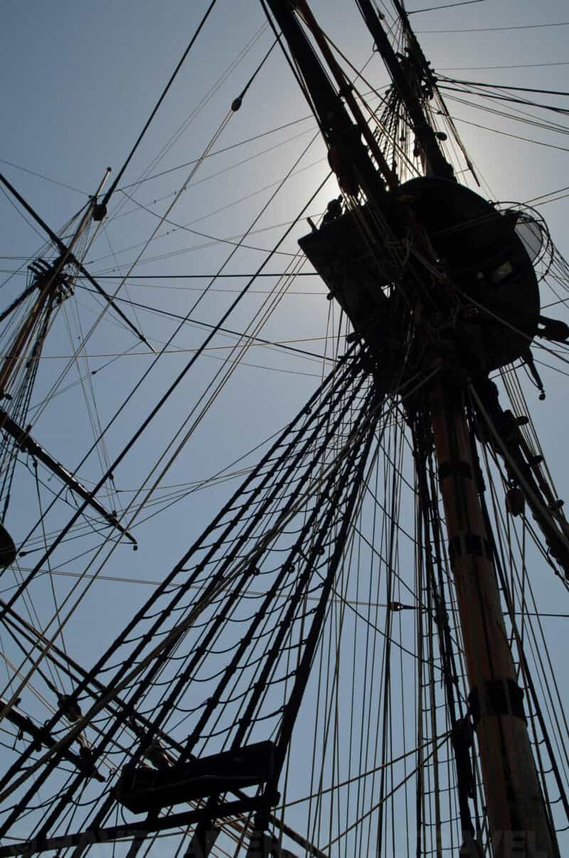 Sun flaring behind a tall ship's mast, rigging and crow's nest silhouetted against blue sky