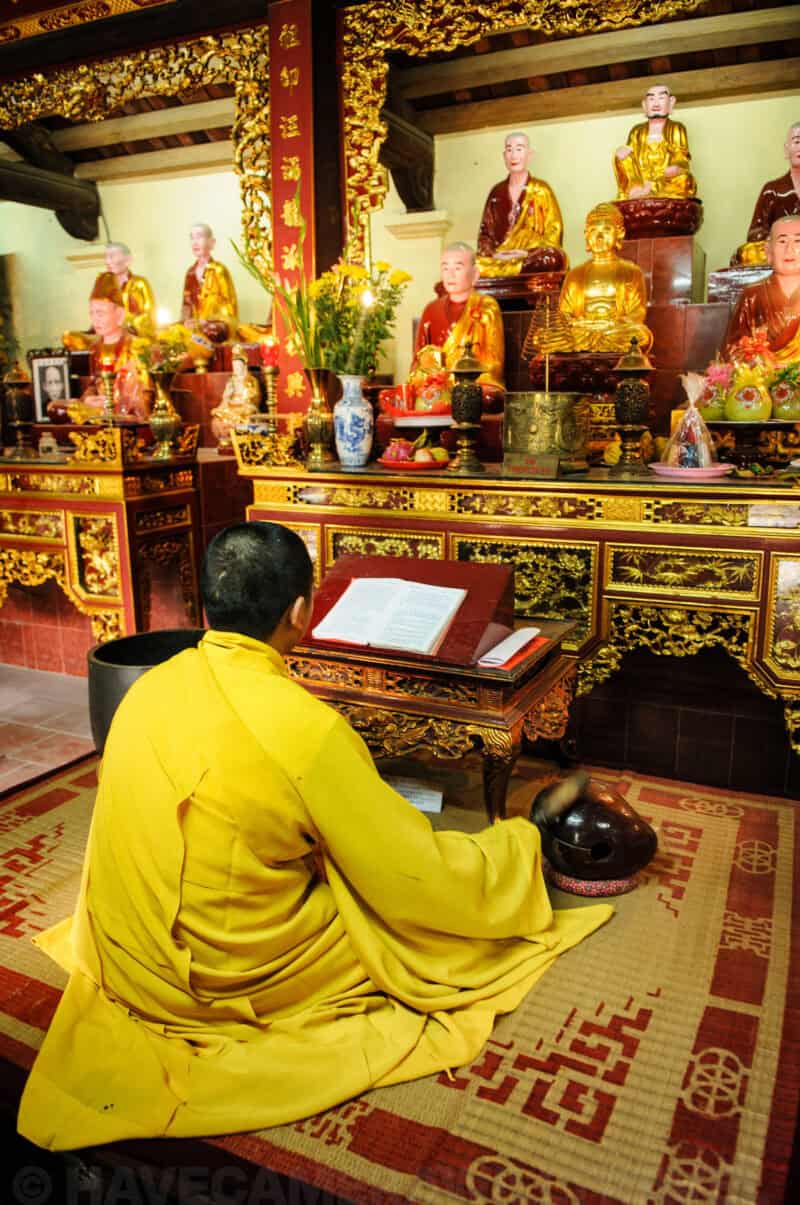 Monk in yellow robes kneeling before gilded Buddha statues at a temple altar in Hanoi