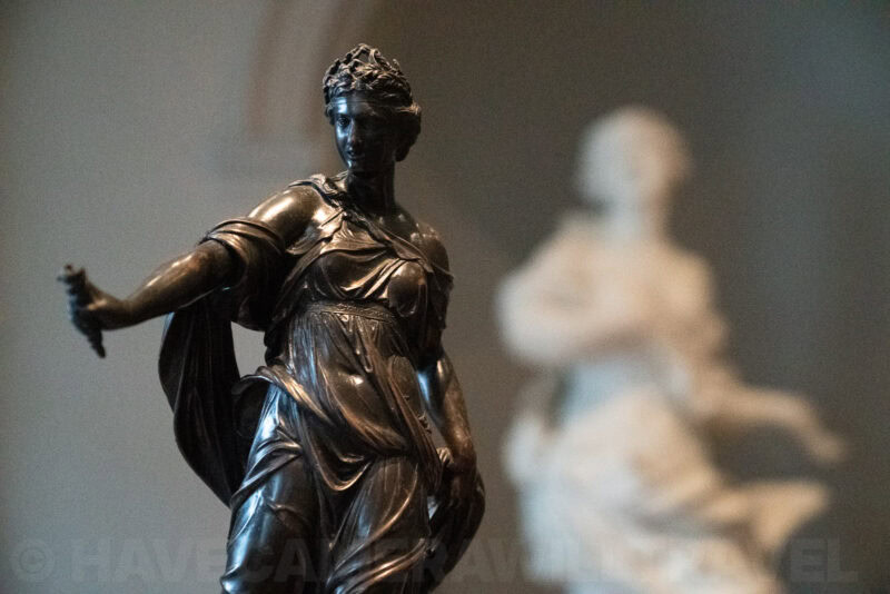 Dark bronze statue of a crowned woman in flowing robes with a blurred marble figure behind