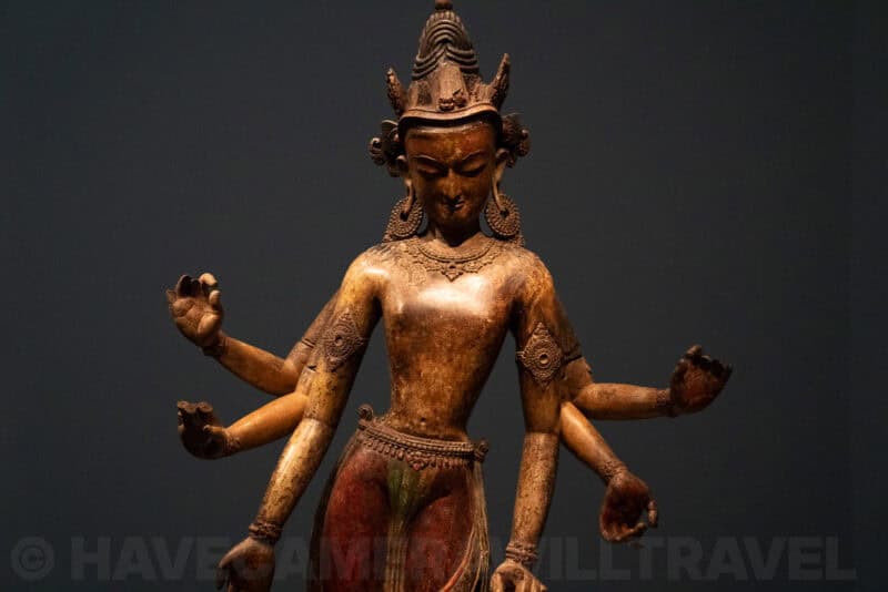 Multi-armed bronze deity statue with ornate crown displayed in a museum gallery