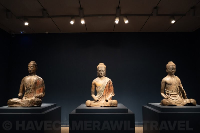 Three seated Buddha statues spotlit against a dark gallery wall in a museum display