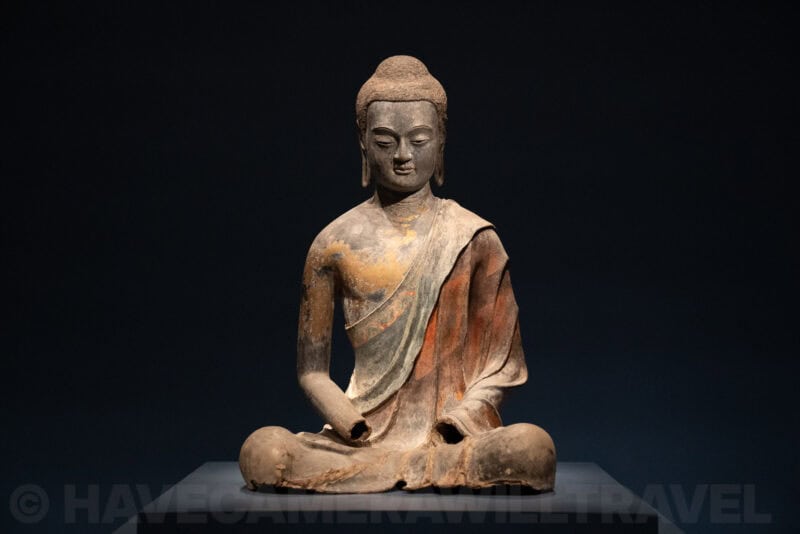 Ancient seated Buddha statue with weathered paint lit against a dark gallery background