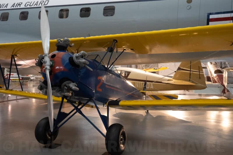 Vintage blue biplane with propeller parked in a hangar beside other historic aircraft