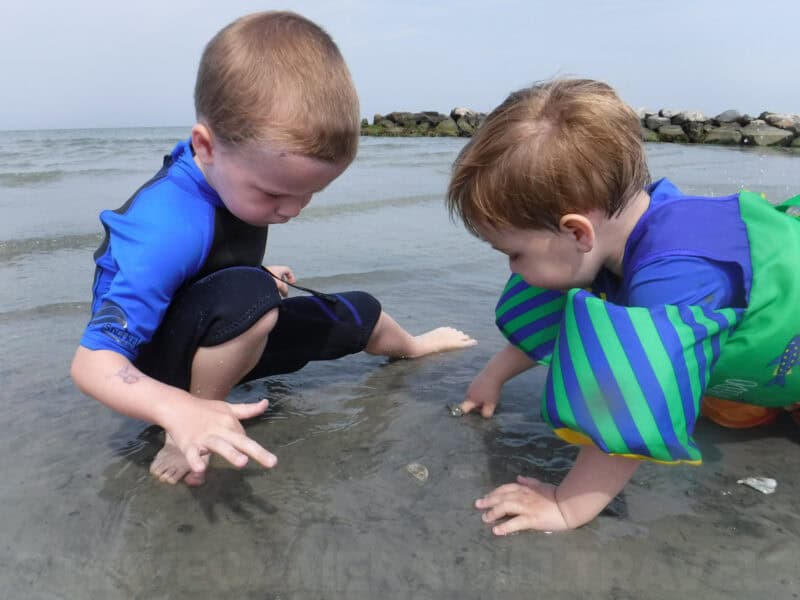 Two young boys in rash guards crouch in shallow surf examining something in the sand