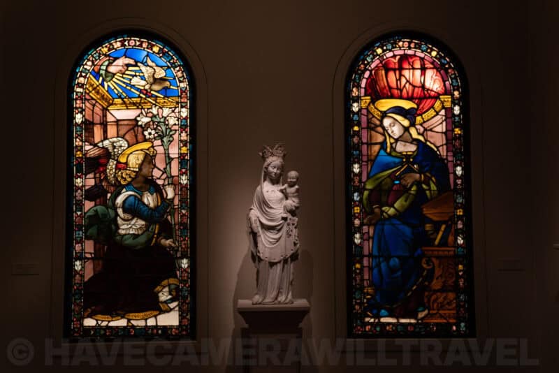 Illuminated stained glass windows flanking a marble statue of Madonna and Child in a museum