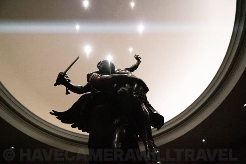 Bronze angel statue with sword seen from below against a domed ceiling with pinpoint lights