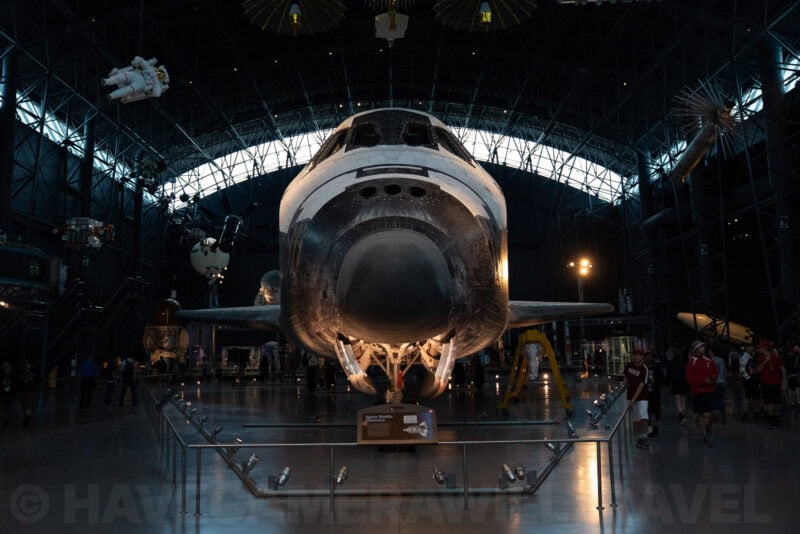 Space shuttle displayed nose-on inside a large museum hangar with visitors walking below