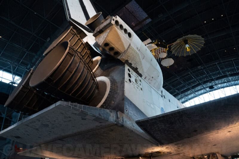 Underside of a space shuttle with exposed engines and heat-shield tiles inside a museum hangar