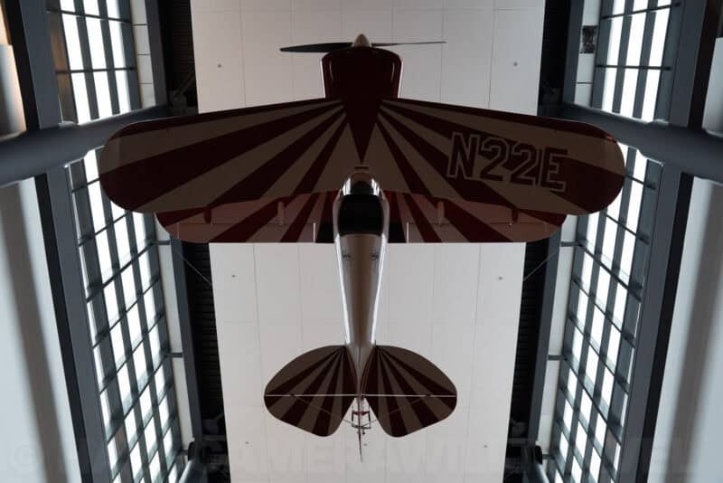 Vintage biplane with red and white striped wings hanging from a museum ceiling, viewed from below