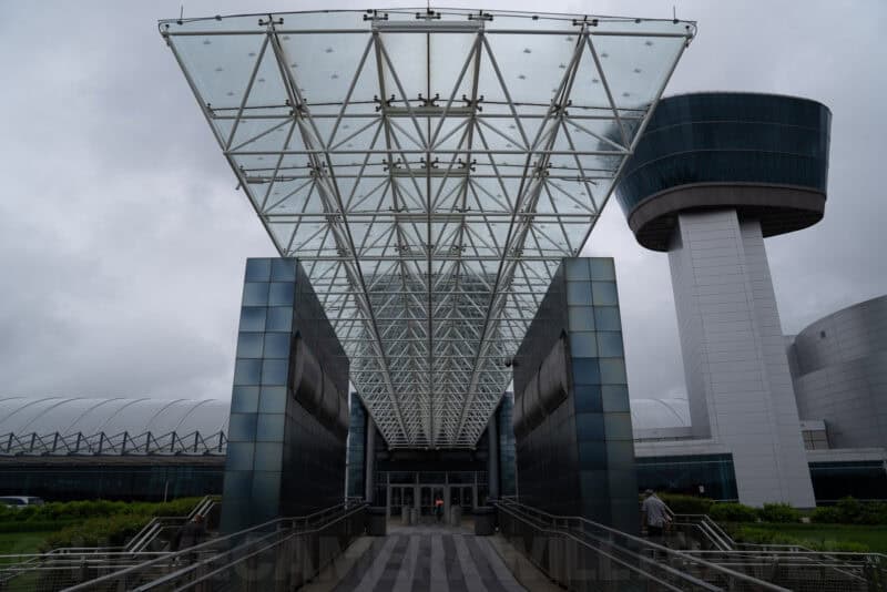 Glass pyramid canopy entrance to an airport terminal with a curved observation tower beyond