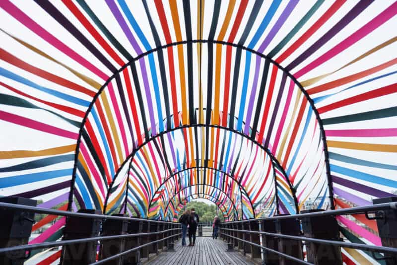 Rainbow-striped tunnel bridge with people walking through its radiating arched canopy outdoors