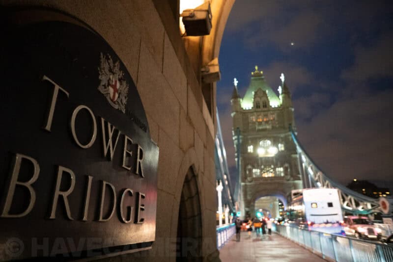 Tower Bridge in London lit at night beside a stone wall plaque reading Tower Bridge