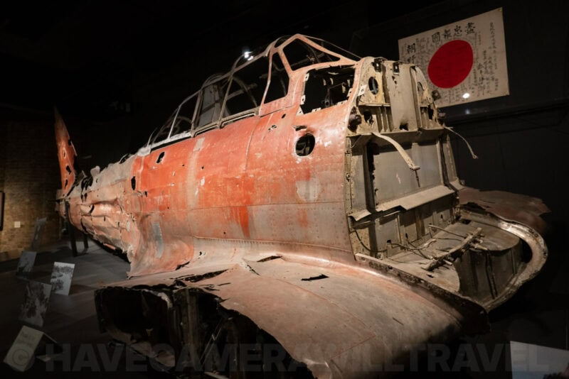 Bullet-riddled wreckage of a WWII Japanese fighter plane fuselage displayed in a dark museum hall