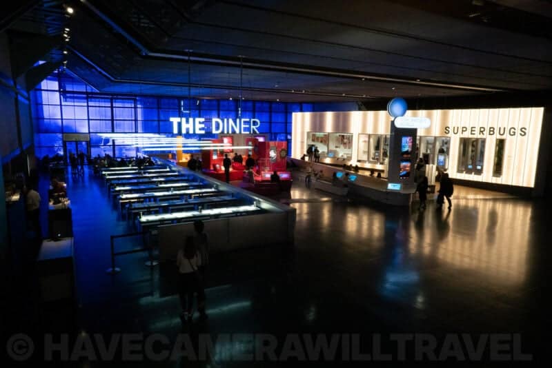 Wide view of a museum hall with THE DINER and SUPERBUGS exhibit signs glowing under blue light