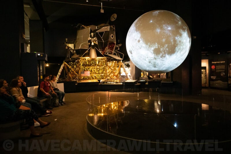 Apollo lunar module replica beside a large illuminated moon sphere in a space museum gallery