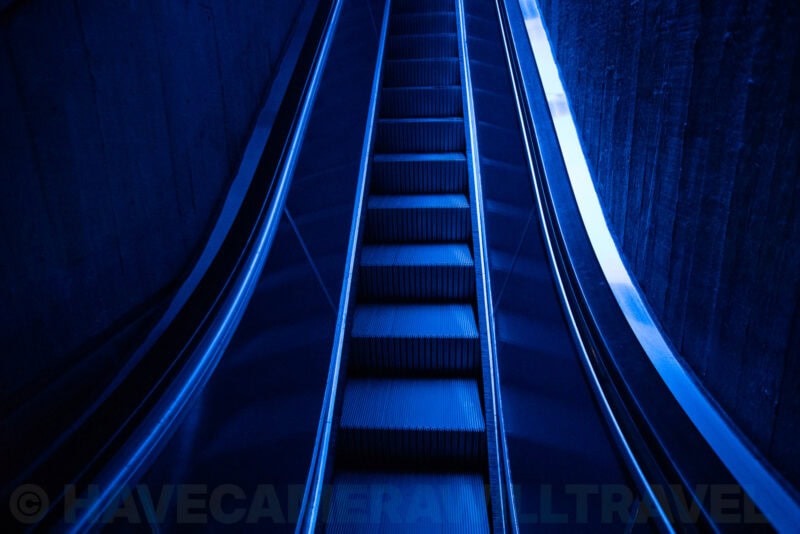 Blue-toned escalator descending steeply between concrete walls in a subway station