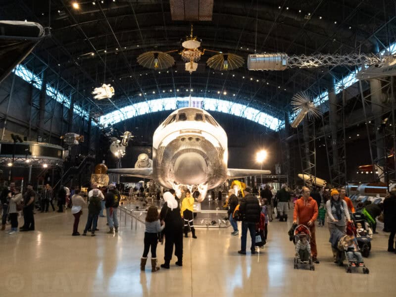 Space Shuttle Discovery displayed nose-on in a large hangar with museum visitors