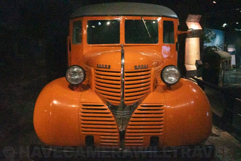 Vintage orange truck with rounded fenders parked in a dark museum exhibit hall