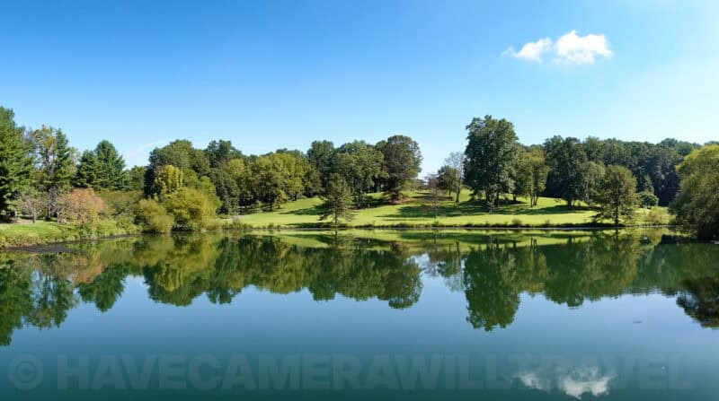 Wide panorama of a calm pond mirroring a tree-lined lawn under blue sky with one white cloud
