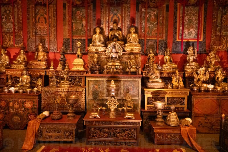 Ornate Buddhist altar with golden statues, ceremonial bowls, and colorful hanging thangkas