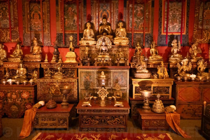 Tibetan Buddhist altar room with gilded statues, offering bowls, and patterned wall hangings