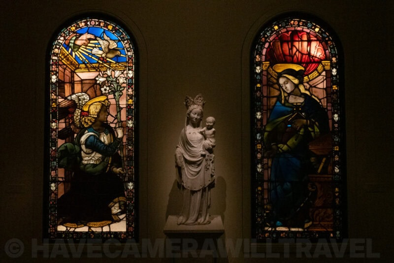 Marble Madonna and Child statue flanked by illuminated stained-glass windows in a museum gallery