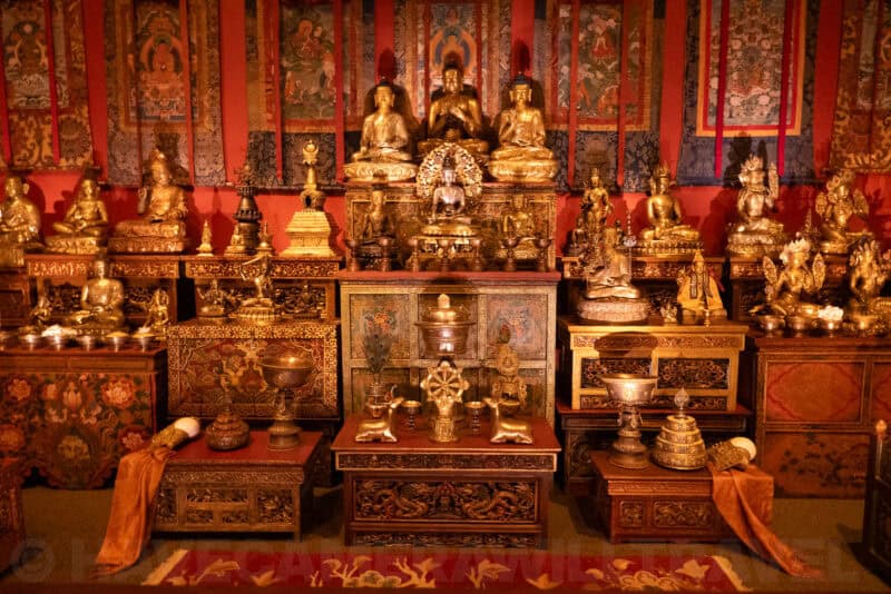 Gilded Buddhist altar with rows of gold statues and hanging thangka scrolls in dim museum lighting