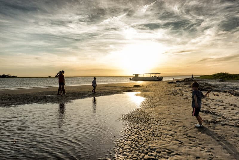 Family walking along a tidal sandbar beach as a tour boat sits near shore at sunset