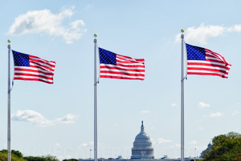 Three American flags waving with the US Capitol dome visible in the distance
