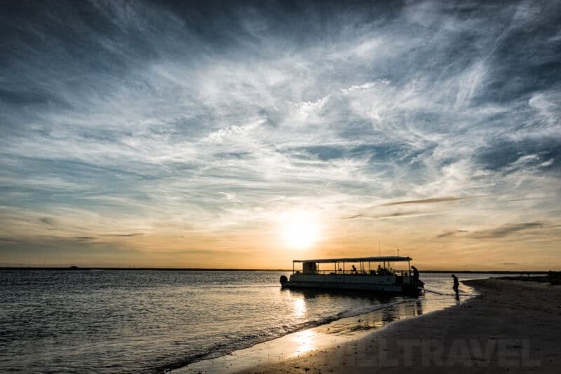 Silhouetted tour boat and person on a beach at sunset with wispy clouds overhead