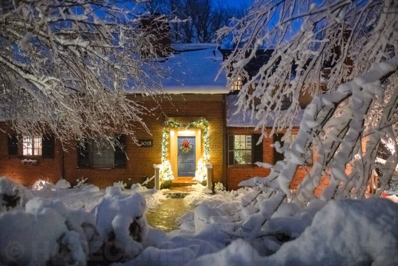 Snow-covered brick house with a wreath and holiday lights glowing at dusk