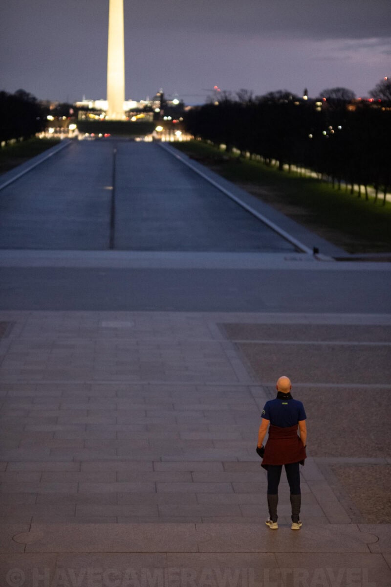 Man standing before the Reflecting Pool with the Washington Monument glowing in the distance at dusk