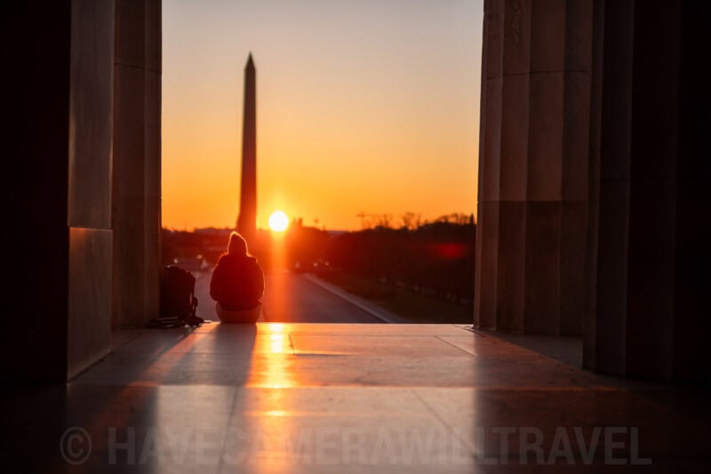 Person sitting between the columns of the Lincoln Memorial facing the sunrise over the Washington Monument