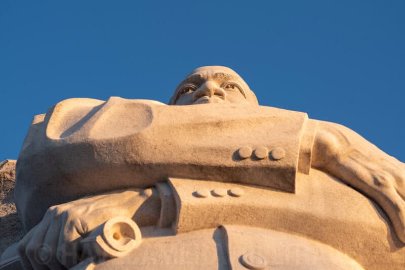 Close-up of a stone Mount Rushmore-style sculpted figure lit warmly against a clear blue sky