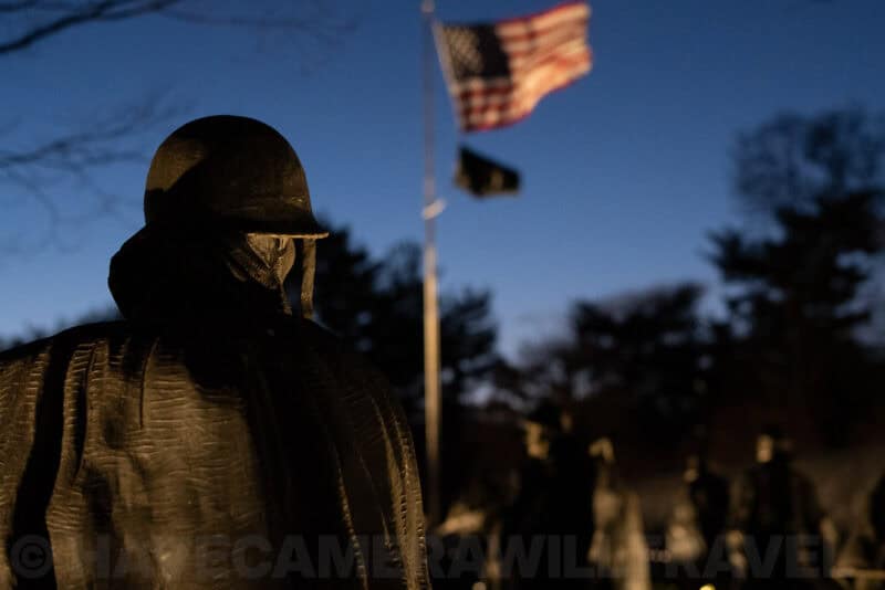 Silhouetted statue at the Korean War Veterans Memorial with an American flag waving against dusk sky