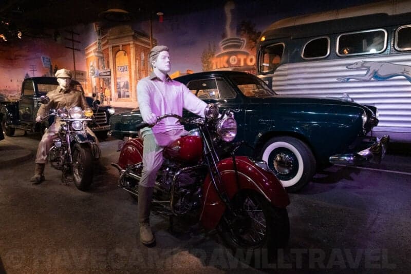 Museum diorama of mannequins on vintage motorcycles beside classic cars, Fujifilm X-T5 ISO 12800 sample shot