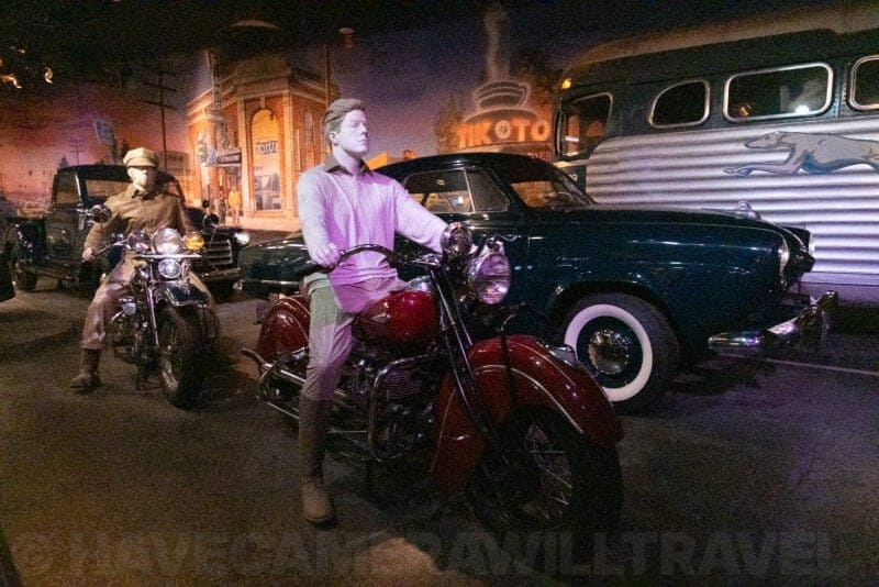 Museum diorama with mannequins on a vintage motorcycle beside classic cars and a bus