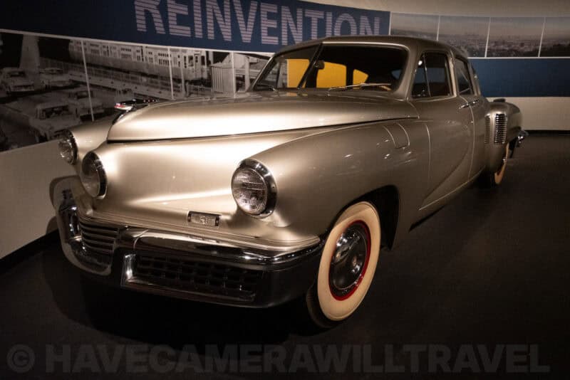 Silver vintage Tucker automobile parked in a museum gallery beneath exhibit signage