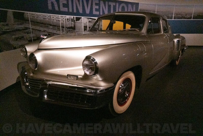Vintage Tucker car with whitewall tires shown in dim museum exhibit lighting