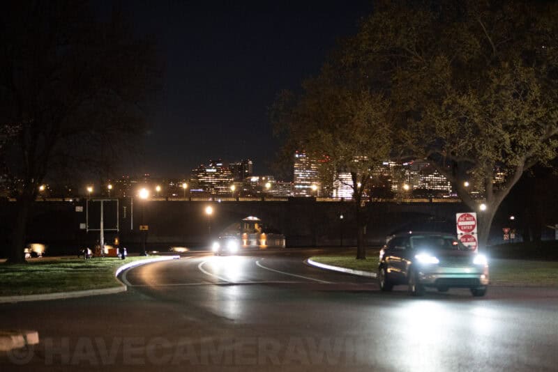 Cars with headlights on a curving nighttime road with a city skyline glowing in the background