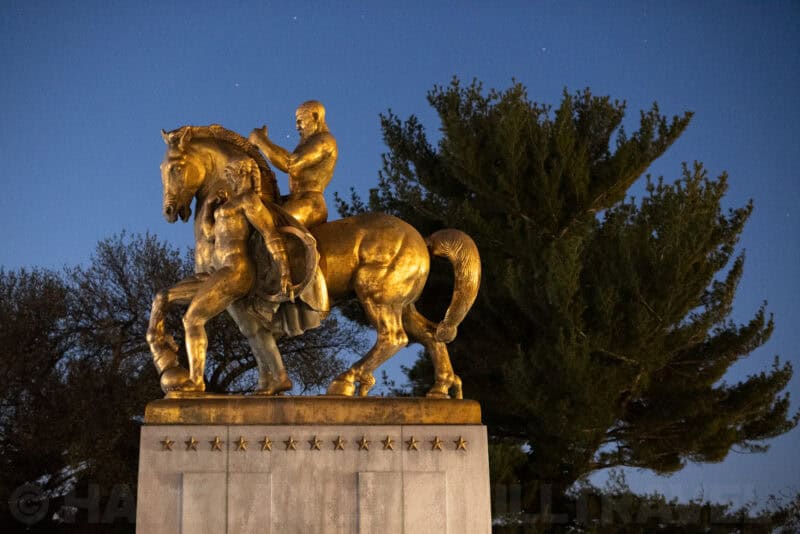 Golden equestrian statue with rider and horse illuminated at dusk with trees behind