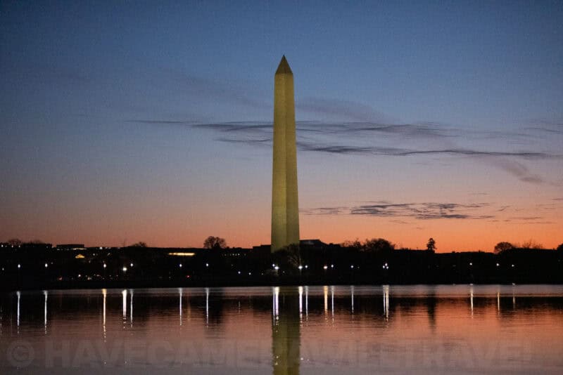 Washington Monument in deepening blue dusk with distant city lights along the water