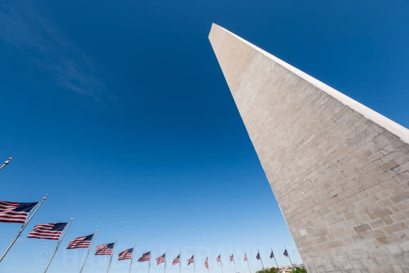 Washington Monument's pointed top and stonework rising against a deep blue sky with flags below