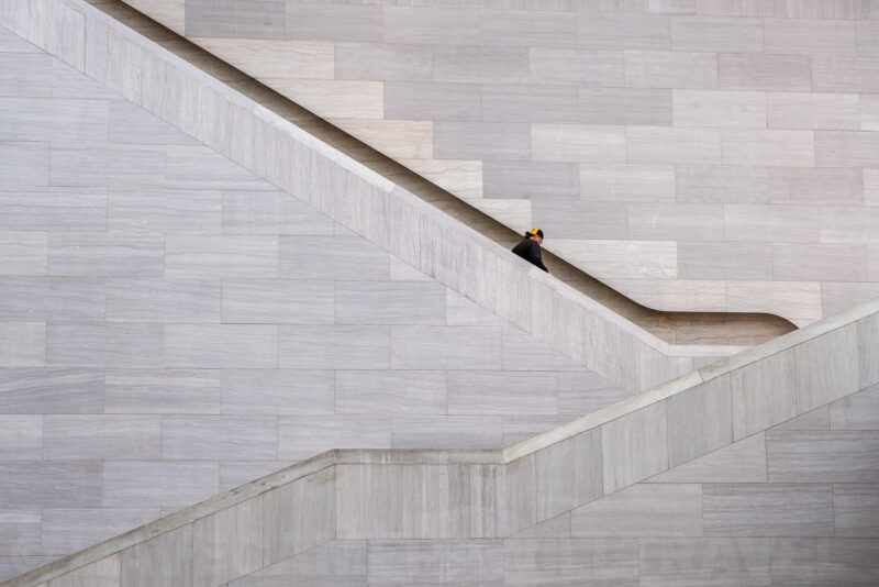 Lone figure walking up a diagonal marble staircase against a minimalist geometric wall