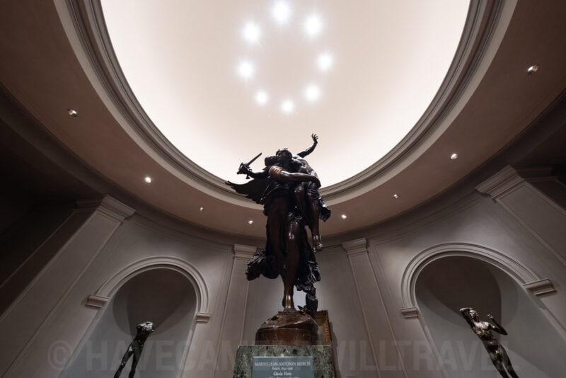 Bronze sculpture of intertwined figures beneath a domed skylight in a museum gallery