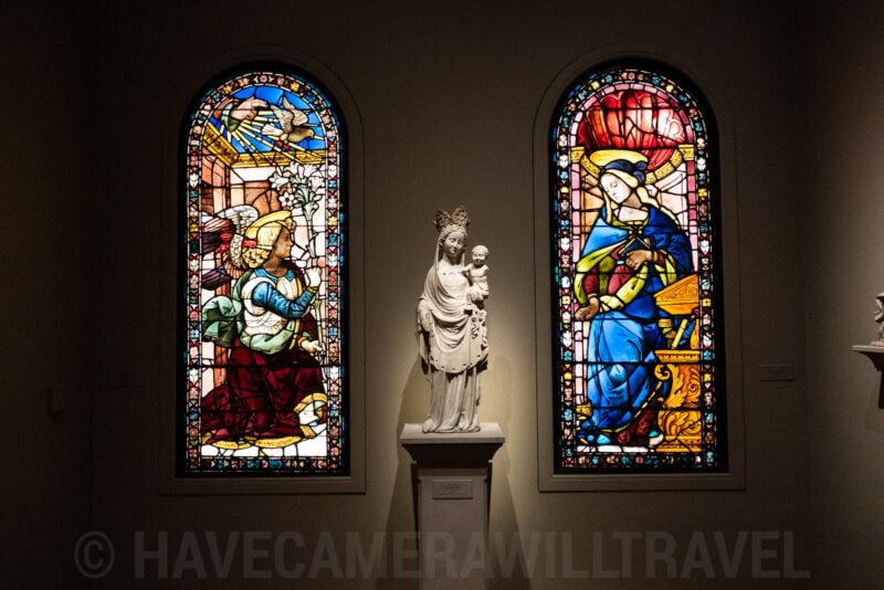 Stained glass windows flanking a marble Madonna and Child statue in a museum gallery
