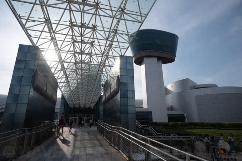 Glass-trussed walkway leading toward a museum building with a rounded observation tower